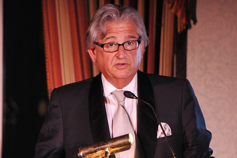 A man with silver hair dressed in a suit speaks at a lectern.